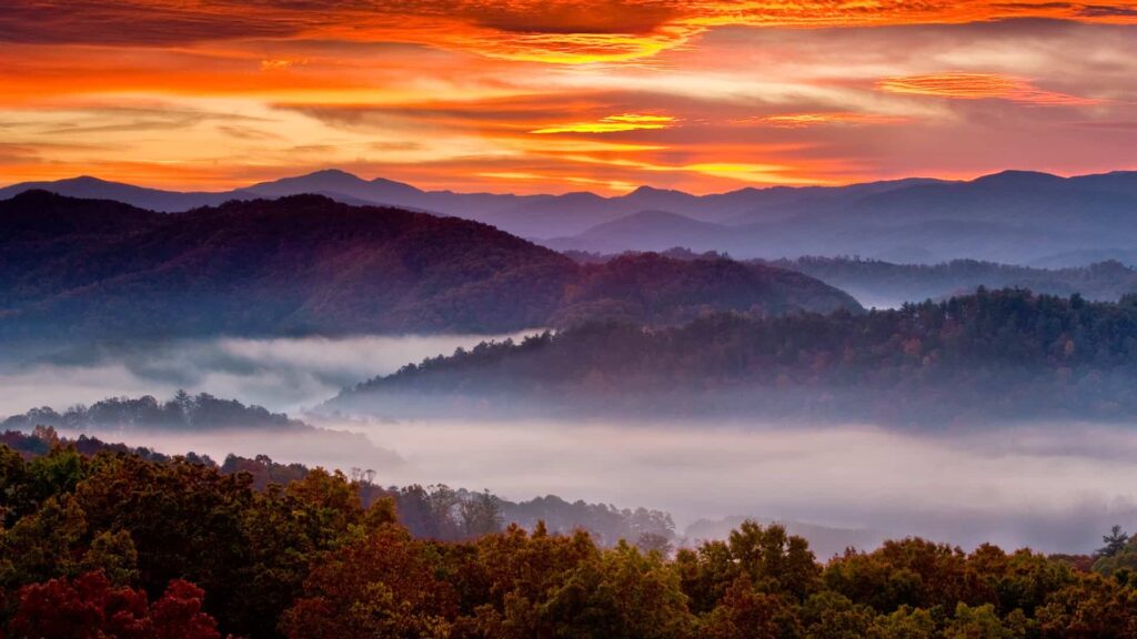 Smoky Mountain National Park Information: Scenic view of Smoky Mountain National Park at sunset. The sky is a vibrant orange-red hue with smoky clouds scattered throughout. In the foreground, the mountain peaks are visible with wispy smoke rising between them, adding to the picturesque landscape.