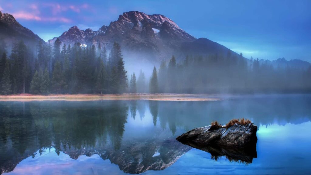 Serene landscape of Grand Teton National Park: Majestic mountain range and tranquil lake with misty morning ambiance and dense coniferous shoreline.